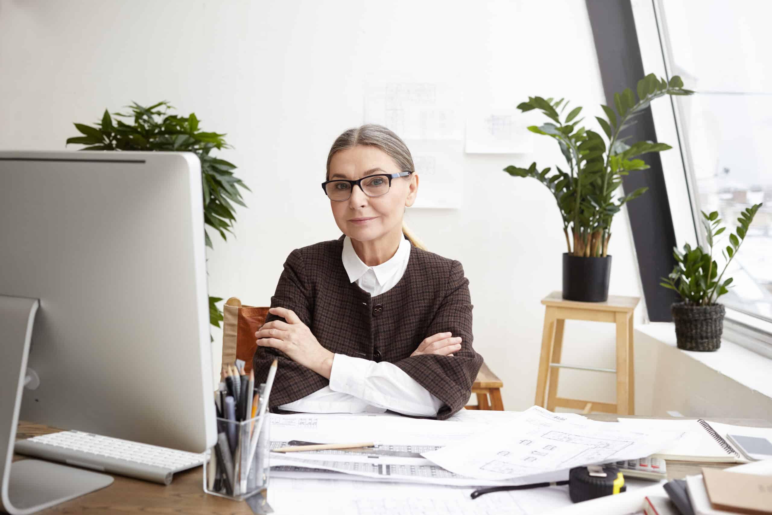 Picture of beautiful gray haired senior Caucasian woman architect in spectacles smiling at camera and keeping arms folded, having rest after she finished drawing of huge architectural project Maximizando o Potencial da Geração Madura: Estratégias para o Engajamento Efetivo no Mercado de Trabalho