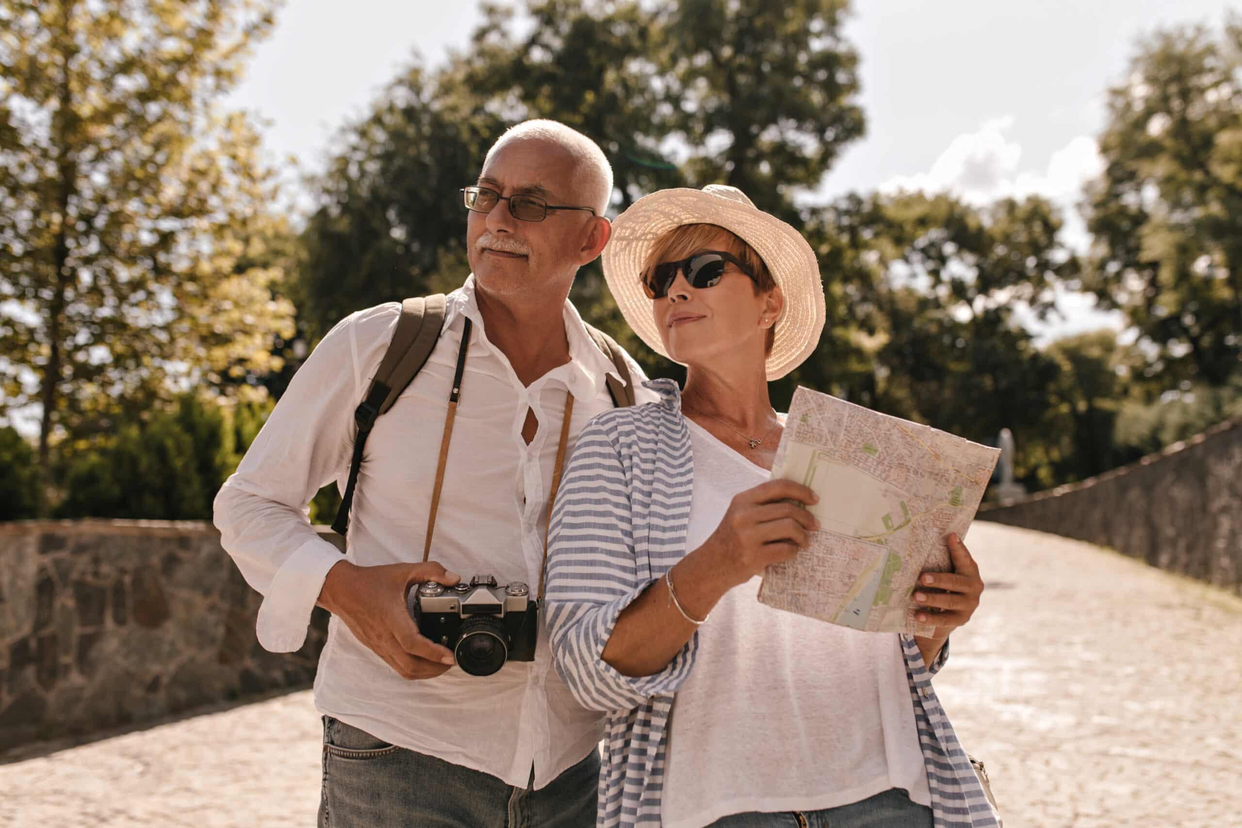 Short haired lady in hat, cool sunglasses and striped blue clothes holding map and posing with man in glasses and white shirt with camera in park. Explorando a Economia Prateada: O Potencial Crescente do Mercado 50+